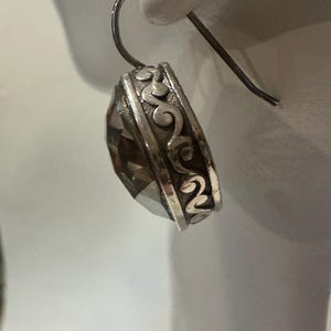 Peut inclure: Boucle d'oreille en argent avec une grosse pierre pr&eacute;cieuse fum&eacute;e et facett&eacute;e. La boucle d'oreille pr&eacute;sente un motif orn&eacute; de volutes autour de la pierre pr&eacute;cieuse et une simple fermeture &agrave; crochet. La boucle d'oreille est pr&eacute;sent&eacute;e sur fond blanc.