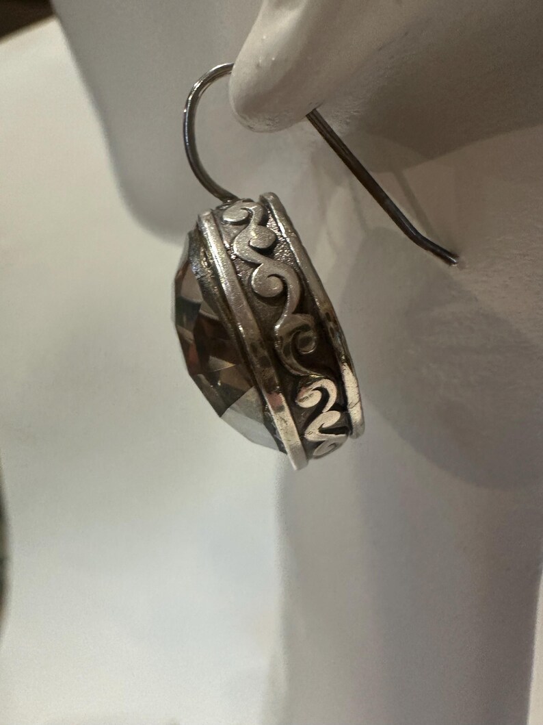 Peut inclure: Boucle d'oreille en argent avec une grosse pierre pr&eacute;cieuse fum&eacute;e et facett&eacute;e. La boucle d'oreille pr&eacute;sente un motif orn&eacute; de volutes autour de la pierre pr&eacute;cieuse et une simple fermeture &agrave; crochet. La boucle d'oreille est pr&eacute;sent&eacute;e sur fond blanc.