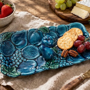 May include: A teal ceramic serving platter with a fruit design, holding crackers, grapes, and pecans. A bowl of strawberries sits nearby. The platter's design includes grapes, plums, and leaves. A cheese board and knife are also present.