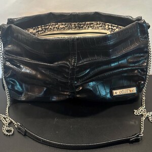 May include: Black faux-leather shoulder bag with a crocodile texture. The bag has a silver chain strap and a small metal tag that reads "REBECCA". The interior lining features a leopard print.