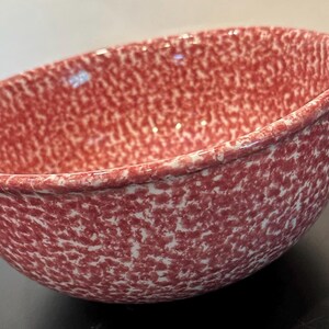 May include: A round ceramic mixing bowl featuring a speckled pattern of deep red and white. The bowl has a smooth, glossy finish and includes a small, integrated pouring spout on the upper rim, designed for controlled pouring of liquid ingredients.