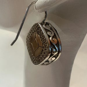 Peut inclure: Boucle d'oreille pendante argent&eacute;e avec un motif g&eacute;om&eacute;trique. La boucle d'oreille pr&eacute;sente une forme centrale dor&eacute;e &agrave; facettes multiples, entour&eacute;e d'un cadre argent&eacute; avec une bordure pointill&eacute;e et des volutes d&eacute;coratives. La boucle est suspendue &agrave; un crochet simple.