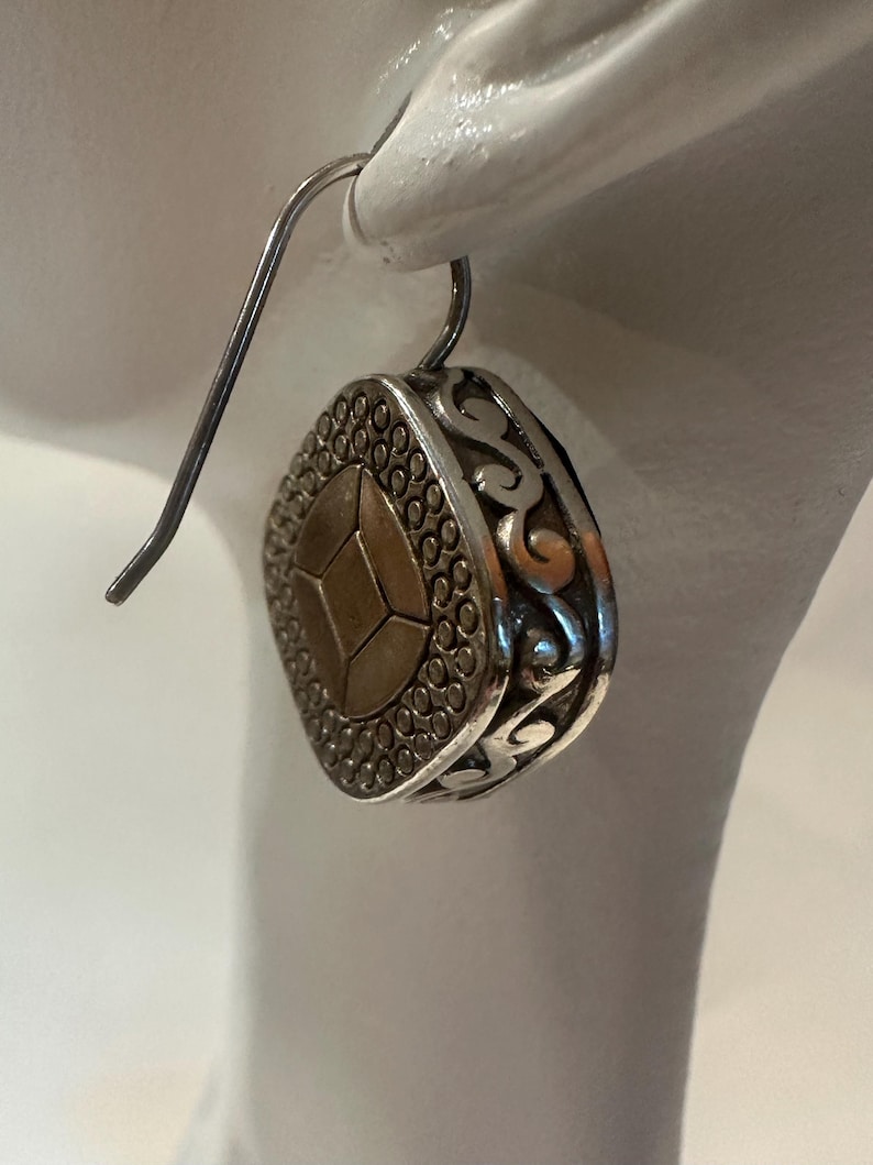 Peut inclure: Boucle d'oreille pendante argent&eacute;e avec un motif g&eacute;om&eacute;trique. La boucle d'oreille pr&eacute;sente une forme centrale dor&eacute;e &agrave; facettes multiples, entour&eacute;e d'un cadre argent&eacute; avec une bordure pointill&eacute;e et des volutes d&eacute;coratives. La boucle est suspendue &agrave; un crochet simple.