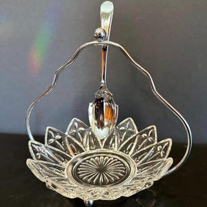 May include: A clear glass dish with a flower-like design, held by a silver-colored metal stand. A small silver spoon hangs above the dish. The dish is approximately 5 inches in diameter.