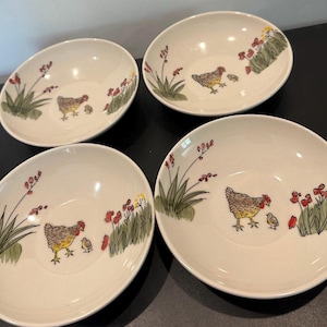 May include: Four round, off-white ceramic bowls, each featuring a hand-painted illustration of a hen and a chick surrounded by green grass, red flowers, and yellow accents. The bowls have a smooth, glossy finish and a simple, shallow design.