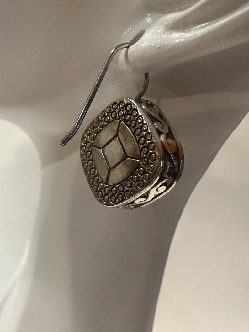 Peut inclure: Boucle d'oreille carr&eacute;e argent&eacute;e avec un motif g&eacute;om&eacute;trique. La boucle d'oreille pr&eacute;sente une forme de losange central entour&eacute;e de petits points. Les bords ont un motif ajour&eacute;. Elle est suspendue &agrave; un crochet d'oreille incurv&eacute;.