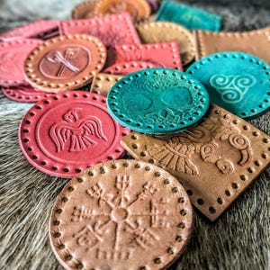 May include: A collection of colorful, embossed leather pieces in various shapes, including circles and squares. The pieces feature intricate designs, such as a tree of life, a raven, and Viking symbols. Colors include shades of red, teal, and brown.