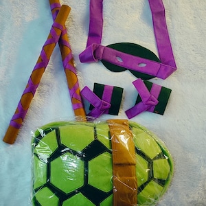 Op de afbeelding: Een Teenage Mutant Ninja Turtles kostuumset met een groene schildpaden schild, twee bruine stokken, een paars masker en twee paarse armbanden.