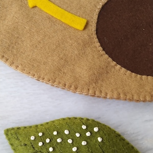 May include: A felt bee with a yellow body and black stripes is flying towards a brown felt circle with a yellow arrow pointing upwards. A green felt leaf with white dots is in the foreground.