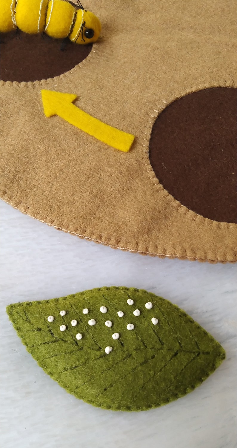 May include: A felt bee with a yellow body and black stripes is flying towards a brown felt circle with a yellow arrow pointing upwards. A green felt leaf with white dots is in the foreground.
