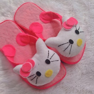 Puede incluir: Un par de pantuflas de fieltro rosa con caras blancas de Hello Kitty. Las pantuflas tienen círculos de fieltro rosa en las tiras y las caras de Hello Kitty tienen ojos negros, narices amarillas y lazos rosas.