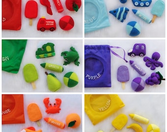 May include: A set of felt color matching bags with various felt objects, including a red fire truck, a green frog, a purple octopus, and a yellow school bus. Each bag has a color name on it, such as "Red", "Green", "Purple", and "Yellow".