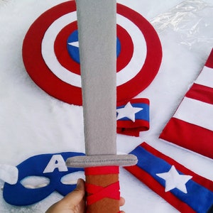 May include: A gray felt sword with a brown and red handle. The sword is part of a Captain America costume set, which also includes a red, white, and blue shield, a blue mask with a white 'A', and two red and blue arm bands with white stars.