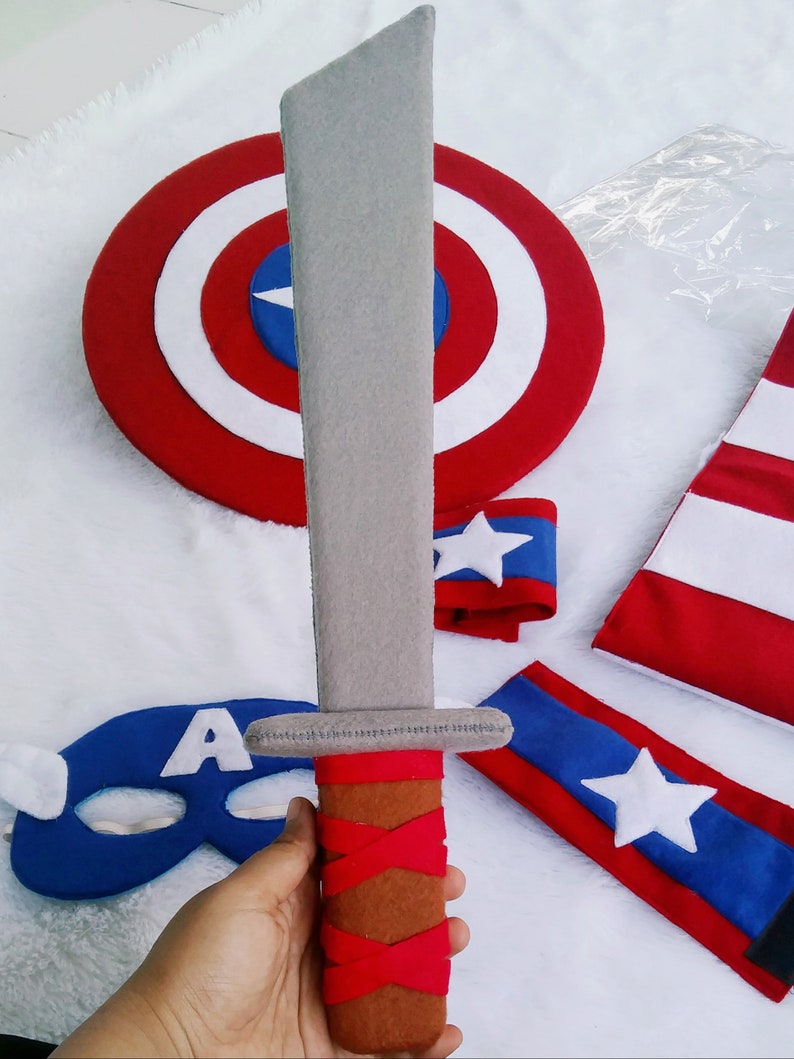 May include: A gray felt sword with a brown and red handle. The sword is part of a Captain America costume set, which also includes a red, white, and blue shield, a blue mask with a white 'A', and two red and blue arm bands with white stars.