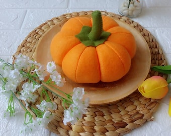May include: A felt orange pumpkin with a green stem, sitting on a woven placemat. The pumpkin is on a wooden plate.