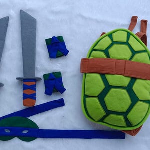 May include: A Teenage Mutant Ninja Turtles costume set with a green turtle shell, a blue mask, two gray swords, and two blue wristbands.