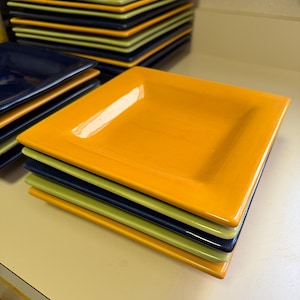 May include: A stack of square ceramic plates in alternating colors of bright orange, navy blue, and lime green. The plates feature a smooth, glossy finish with a slightly raised rim and a flat center, arranged in a neat pile on a flat surface.