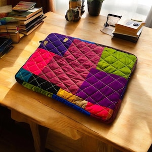 Peut inclure: Une housse d'ordinateur portable en patchwork coloré avec un motif matelassé. La housse est faite de différents tissus dans des tons de rose, violet, vert, bleu et jaune.