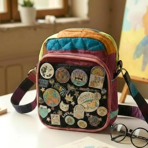 Peut inclure: Un sac bandoulière coloré avec un motif patchwork, orné de nombreuses épingles. Le sac présente un panneau avant noir recouvert de diverses épingles rondes et de formes variées. La sangle est multicolore. Des lunettes rondes et un livre sont sur la table.