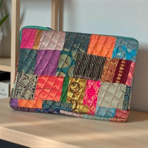 Puede incluir: Una funda para portátil de patchwork con una variedad de telas coloridas en tonos rosa, naranja, verde y morado. La funda tiene un diseño acolchado y una cremallera turquesa. La funda rectangular está sobre una superficie de madera.