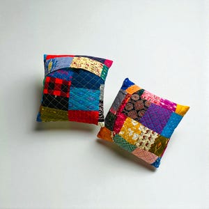 Puede incluir: Dos cojines cuadrados de patchwork con una variedad de cuadrados de tela coloridos. Los cojines tienen un diseño acolchado y están rellenos con un material suave. La paleta de colores incluye rojo, azul, amarillo y verde.