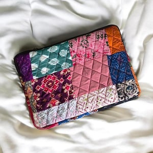 Puede incluir: Una funda rectangular para portátil con un diseño de patchwork acolchado que combina varios patrones de tela en tonos rosa, azul verdoso, morado, naranja y azul. La superficie muestra motivos geométricos, florales y figuras humanas.