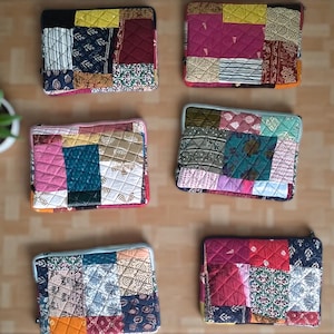 May include: A collection of six colorful patchwork laptop sleeves. Each sleeve features a unique design with various fabric patterns and colors, including blue, pink, yellow, and red. The sleeves are arranged on a light brown surface.