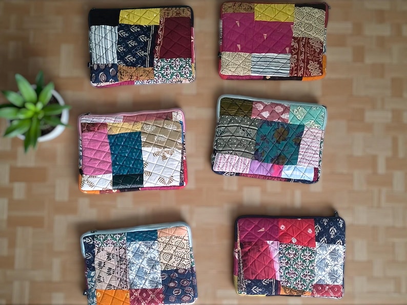 May include: A collection of six colorful patchwork laptop sleeves. Each sleeve features a unique design with various fabric patterns and colors, including blue, pink, yellow, and red. The sleeves are arranged on a light brown surface.