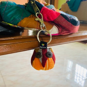 Può includere: Un portachiavi a forma di gufo in tessuto colorato con occhi di bottone marroni e un'etichetta che dice "Upcycle" con un cuore.