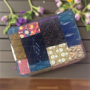 May include: A quilted patchwork zippered pouch featuring a variety of patterned fabric squares in shades of blue, brown, pink, and green. The pouch has a diamond-stitched pattern and a dark blue zipper. The overall design is a mix of textures and colors.