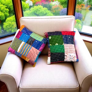 Op de afbeelding: Twee patchwork sierkussens met een verscheidenheid aan kleurrijke stoffen vierkanten liggen op een crèmekleurige fauteuil. De kussens hebben een mix van patronen en texturen, waaronder gewatteerde stiksels en geometrische ontwerpen. Een levendige tuin is zichtbaar door het raam.