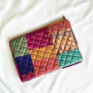 Puede incluir: Una funda rectangular acolchada para portátil con un diseño de patchwork de cuadrados de tela brillante en tonos magenta, oro, verde azulado, morado y naranja. La funda tiene una cremallera negra a lo largo del borde superior y textura.