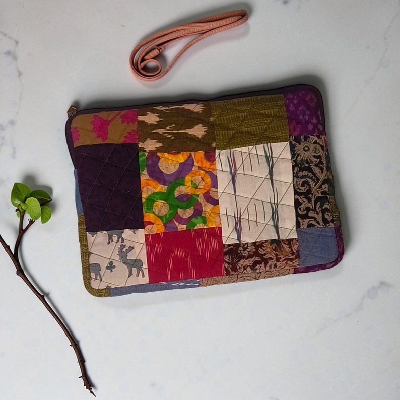May include: A patchwork laptop sleeve with a variety of fabric patterns in shades of purple, green, brown, and red. The sleeve has a zipper closure and a detachable pink strap. The design is a mix of geometric and floral patterns.