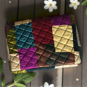 May include: A quilted clutch with a patchwork design. The clutch features a variety of colors, including gold, teal, purple, and red. The fabric has a diamond pattern. The clutch is rectangular and has a zipper closure.