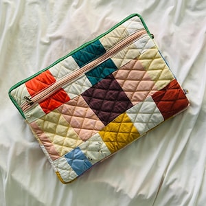 May include: A rectangular quilted laptop sleeve featuring a patchwork design of various solid and patterned fabric squares in shades of orange, teal, yellow, pink, and cream. The sleeve has a green fabric border and a light pink zippered pocket.