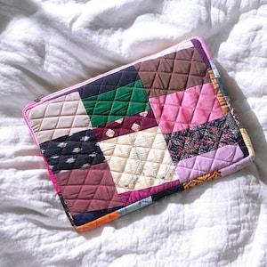 May include: A rectangular quilted laptop sleeve featuring a patchwork design of various fabric squares in shades of brown, green, pink, cream, black, and orange. The sleeve has a pink zip closure along the top edge and a diamond-stitched pattern.
