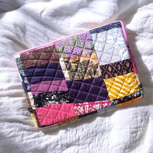 Puede incluir: Una funda acolchada rectangular para portátil con un diseño de patchwork de varios patrones de tela en tonos morado, rosa, amarillo, marrón y negro. La funda tiene una cremallera rosa brillante en el borde superior y una pequeña etiqueta negra.