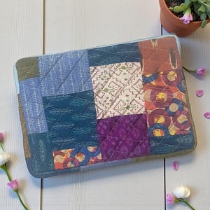 May include: A patchwork laptop sleeve featuring a variety of patterned fabrics in shades of blue, green, purple, and orange. The sleeve has a quilted design and a light blue zipper. The sleeve is rectangular and appears to be approximately 14 inches long.