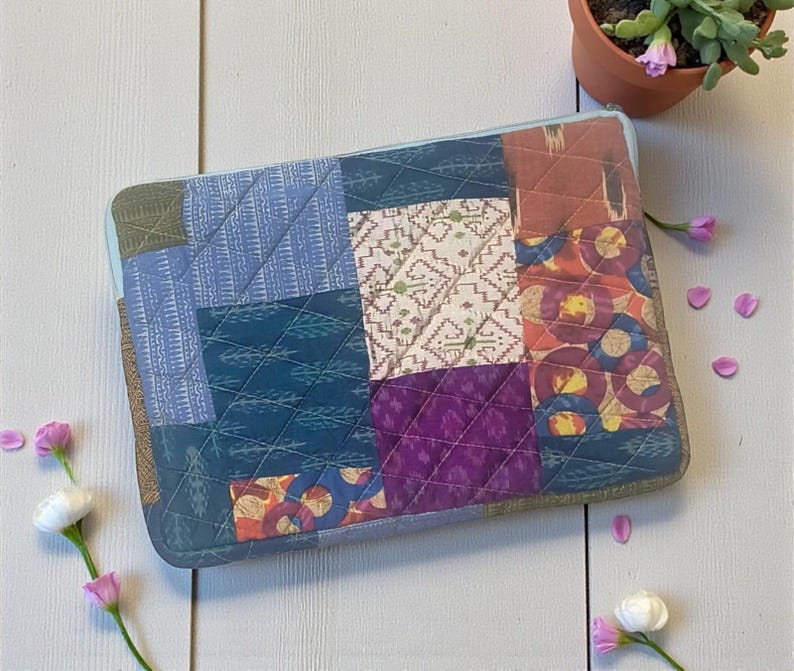 May include: A patchwork laptop sleeve featuring a variety of patterned fabrics in shades of blue, green, purple, and orange. The sleeve has a quilted design and a light blue zipper. The sleeve is rectangular and appears to be approximately 14 inches long.