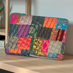 May include: A patchwork laptop sleeve with a teal zipper. The sleeve is made of various colorful fabric squares, including pink, orange, purple, and gold, stitched together in a quilted pattern. The sleeve is rectangular and sits on a wooden surface.