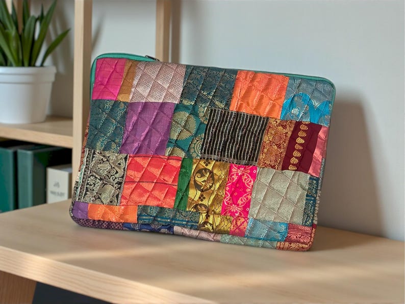 May include: A patchwork laptop sleeve with a teal zipper. The sleeve is made of various colorful fabric squares, including pink, orange, purple, and gold, stitched together in a quilted pattern. The sleeve is rectangular and sits on a wooden surface.