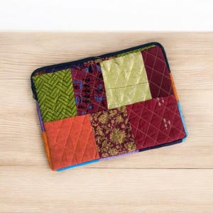 May include: A patchwork laptop sleeve in various fabrics, including green, orange, burgundy, and gold. The sleeve features a quilted design and a dark blue zipper. The sleeve is approximately 33 cm wide and 25 cm tall.