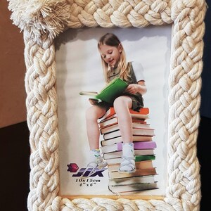 Puede incluir: Un marco de fotos de cuerda blanca con un diseño trenzado y un pompón blanco en la esquina superior. El marco contiene una foto de una niña sentada en una pila de libros. La foto está etiquetada "10x15cm 4"x6""