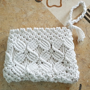 May include: A white macrame clutch bag with a tassel. The bag has a woven design with a geometric pattern.