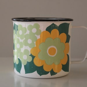 Puede incluir: Taza de esmalte blanco con borde negro y asa blanca. La taza está decorada con un estampado floral en tonos verdes y amarillos. Las flores están sobre un fondo blanco.