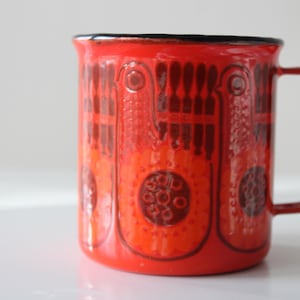 Puede incluir: Una taza de esmalte roja con un borde y un asa negros. La taza tiene un patrón geométrico en negro y naranja.