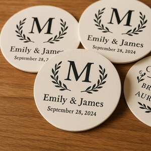 Puede incluir: Un conjunto de posavasos redondos con fondo crema. Cada uno presenta una letra "M" negra rodeada por una corona de laurel. El texto "Emily & James" y "September 28, 2024" están impresos debajo. Sobre una superficie de madera.