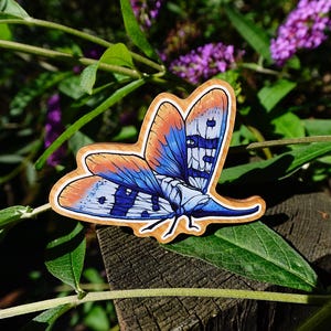 Könnte beinhalten: Ein hölzerner Schmetterlings-Ansteckpin mit orangefarbenen, blauen und weißen Flügeln. Der Schmetterling hat ein detailliertes Design und ist vor einem natürlichen Hintergrund aus grünen Blättern und lila Blumen platziert. Der Pin ist aus Holz.
