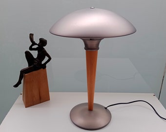 Puede incluir: Una lámpara de mesa plateada con una pantalla ancha y redonda de plata y una base de madera. La lámpara está sobre una superficie blanca.