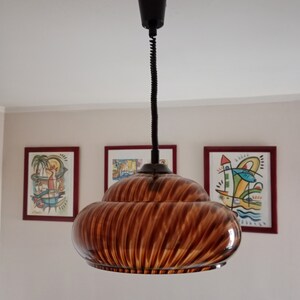 May include: A brown glass pendant light with a swirling pattern hangs from a black cord. The light is suspended from a ceiling in a room with white walls and three framed pictures.